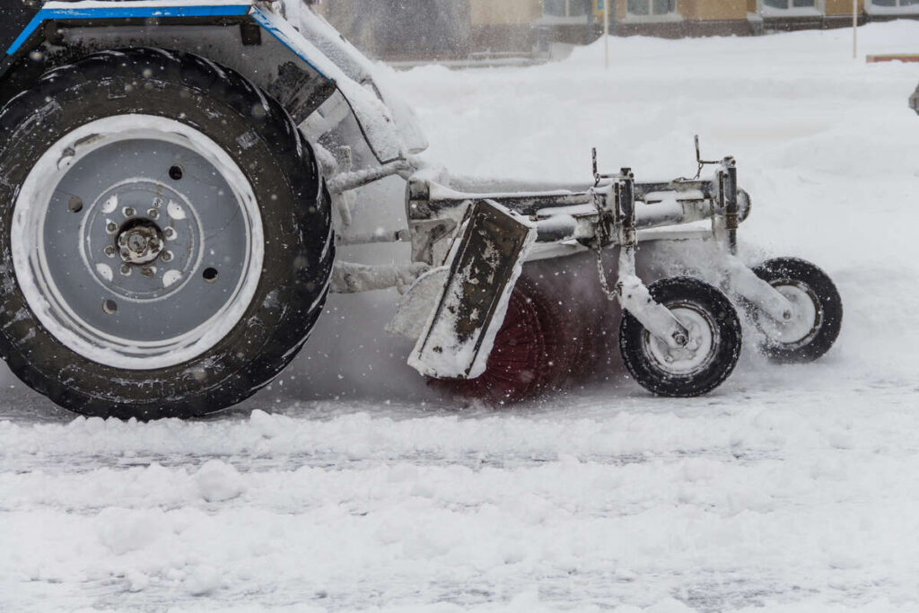 Snow-covered tractor with plow attachment clearing winter roads during a heavy snowfall